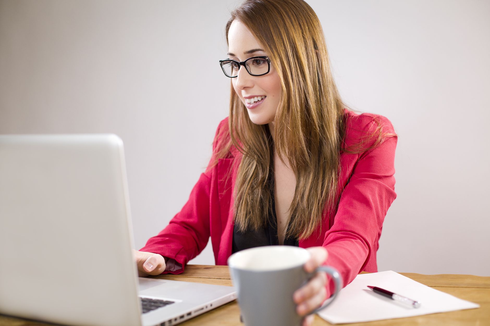 Jeune femme d'affaires souriante en veste rouge travaillant sur son ordinateur portable en tenant une tasse de café.